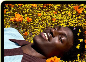 MacBook Neo, kleur Citrus, op het scherm een lachende persoon die in een zonnig bloemenveld ligt